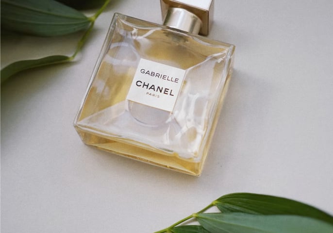 chanel gabrielle perfume laying on a white surface with green leaves at the top left and bottom right of it